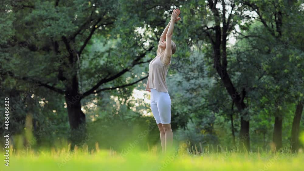 Young woman doing yoga practice or fitness while standing in green summer park spbi. 4k video Attractive athletic female does asana or pose and raises hands to sky, stands on grass and stretches her Young woman doing yoga practice or fitness while standing in green summer park spbi. 4k video Attractive athletic female does asana or pose and raises hands to sky, stands on grass and stretches her
