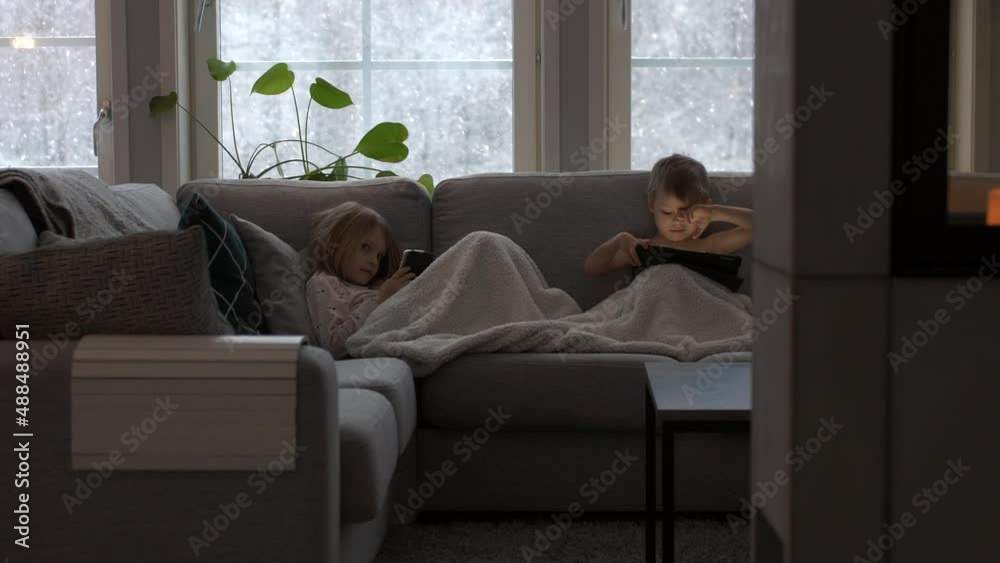 Children sit on couch with pad and phone, girl smiles into camera