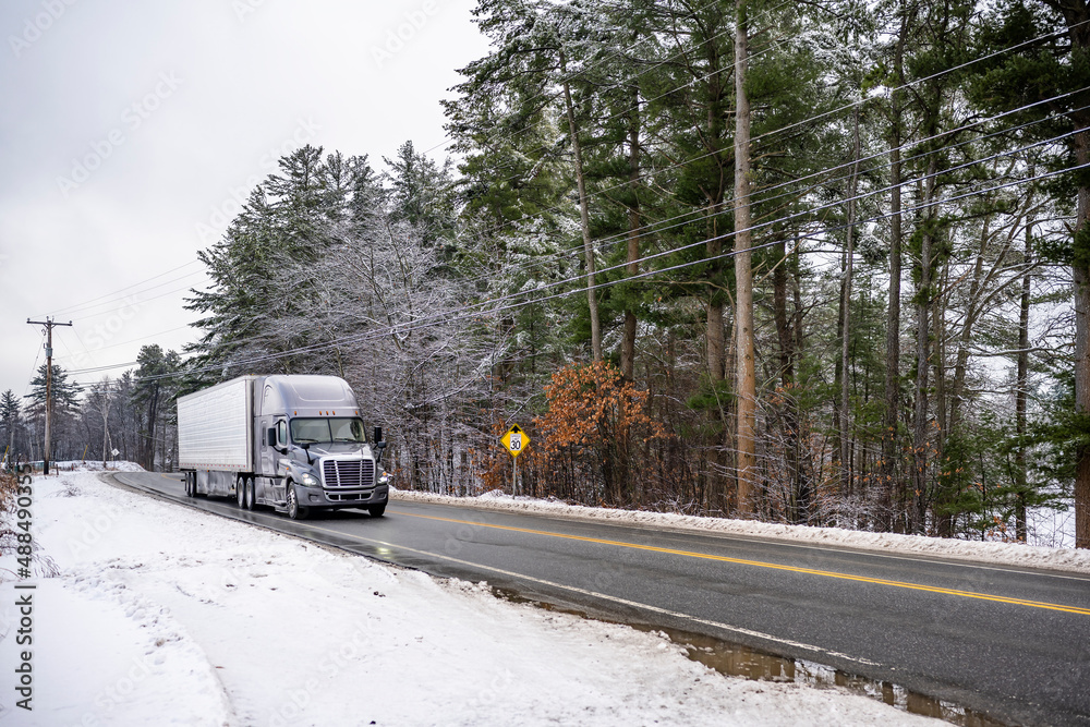 Gray big rig semi truck transporting cargo in refrigerator semi trailer running on the local winter road with snow and trees on the sides