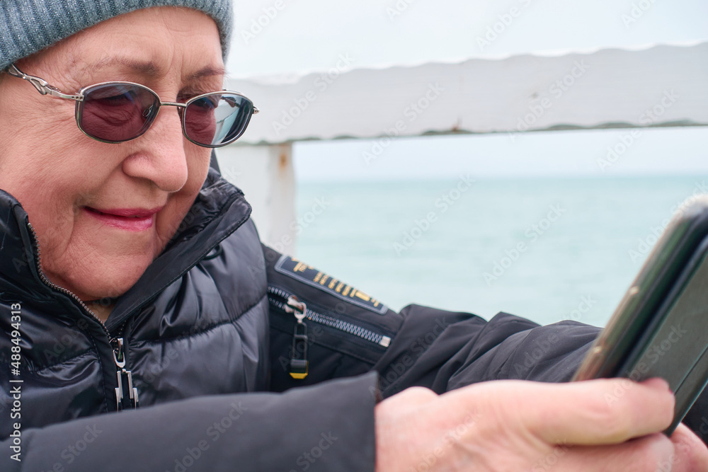An elderly woman in glasses, a hat and winter clothes sits at a table and writes a message on the phone. Selective focus.