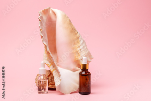 Transparent and brown cosmetic bottles with dropper on a pink background with decor of strombus gigas sea shell. Copy space. The concept of an advertising template for beauty products photo