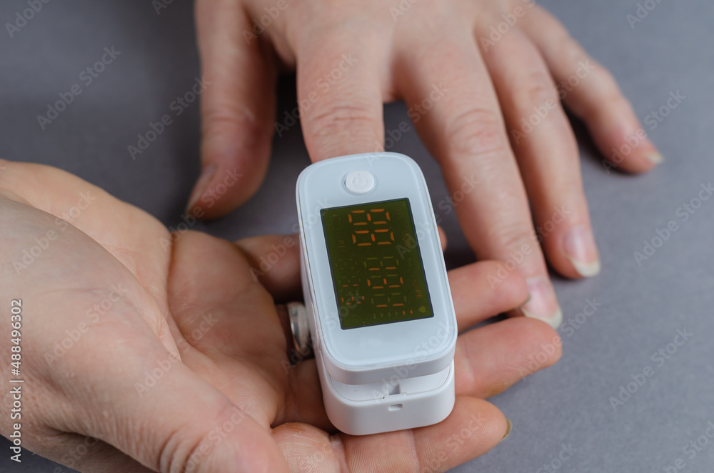 The oximeter is worn on the index finger of a woman's hand. A device for measuring blood oxygen levels and heart beats. Hands of an adult female against a gray background. Indecipherable readings. The oximeter is worn on the index finger of a woman's hand. A device for measuring blood oxygen levels and heart beats. Hands of an adult female against a gray background. Indecipherable readings.