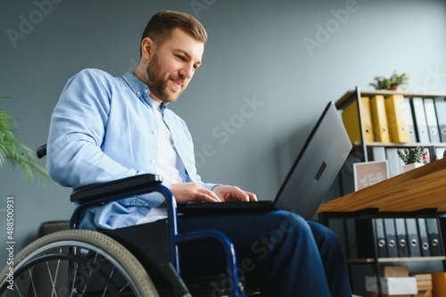 Disabled person in the wheelchair works in the office at the computer. He is smiling and passionate about the workflow.