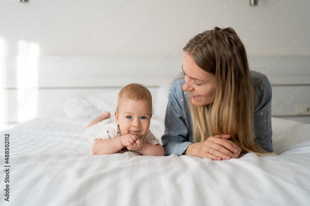 mother with baby six months old in the bedroom. Young happy mum lies next to her adorable baby daughter playing in white bed enjoying sunny morning mother with baby six months old in the bedroom. Young happy mum lies next to her adorable baby daughter playing in white bed enjoying sunny morning