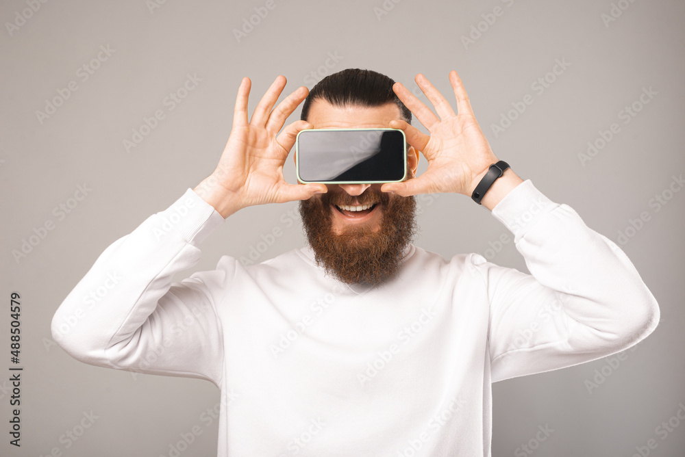 Young bearded man is covering eyes with his smartphone. Studio shot. Young bearded man is covering eyes with his smartphone. Studio shot.