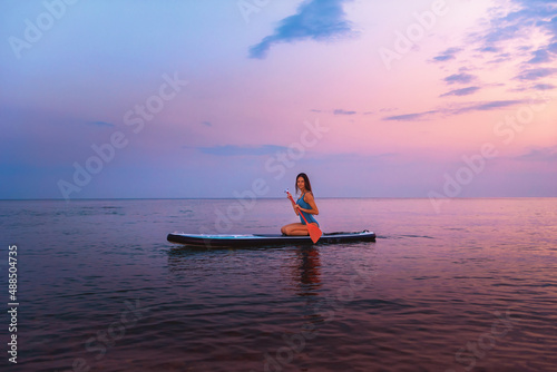 A young smiling woman poses sitting on a sup board with paddle. Sunset in the background. Copy space. Concept of sup boarding