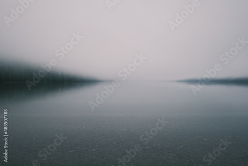 Beautiful mystical landscape of Altai Lake on a foggy morning. Shore with calm clear transparent water and pebbles at the bottom, in the background is a strip of coniferous trees in a foggy haze.