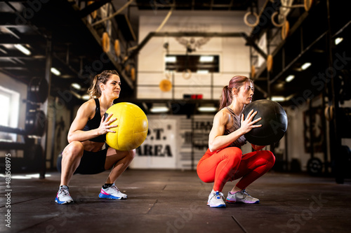 Two woman doing squats while holding heavy medecine balls. Fitness women doing bodyweight exercises using weight ball. People in a row squatting with weighted balls at cross training gym.