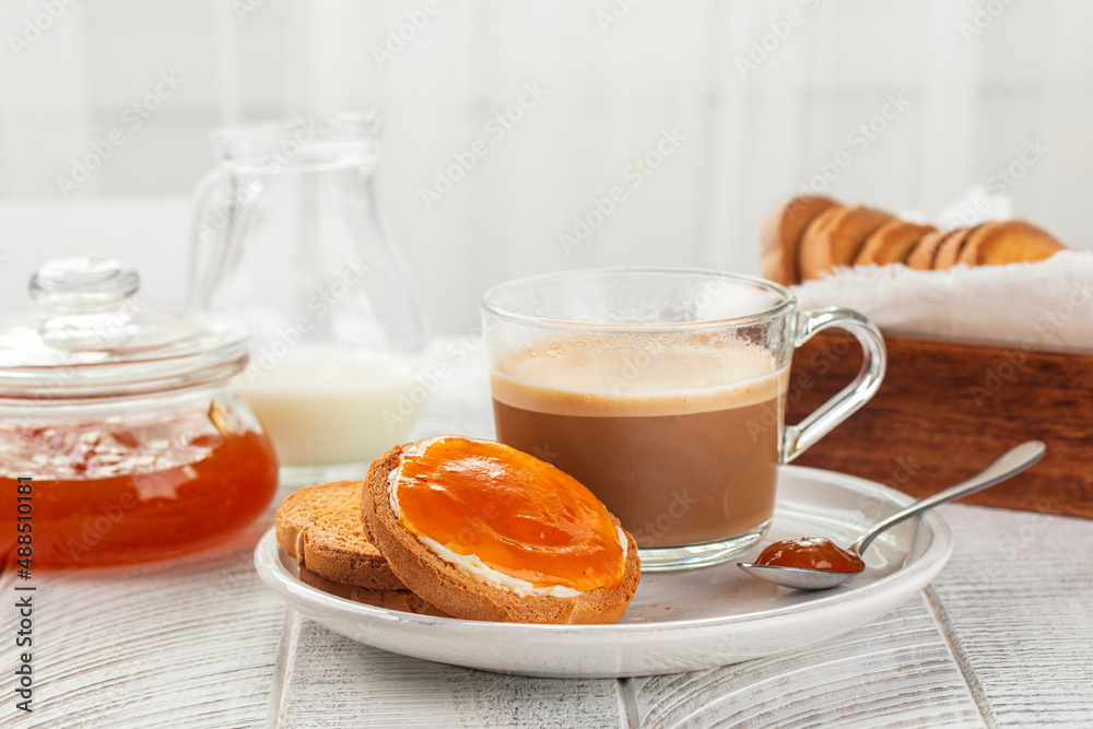 Traditional Italian Breakfast. Apricot Jam and Butter on Rusks or Fette Biscottate, toasted round Bread. Caffe lungo or morning coffee. Milk. White table and background. Copy space. Traditional Italian Breakfast. Apricot Jam and Butter on Rusks or Fette Biscottate, toasted round Bread. Caffe lungo or morning coffee. Milk. White table and background. Copy space.