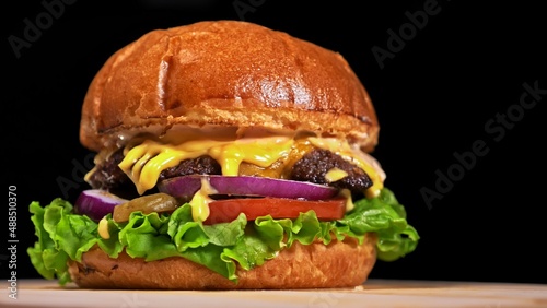 Craft burger is cooking on black background in black food gloves. Consist: sauce, lettuce, tomato, red onion, pickle, cheese, bacon, air bun and marbled meat beef. Not made ideal. Looks real, loving