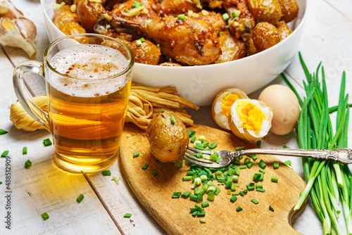 lunch in rural style, tasty food - glass of beer, fried chicken meat and potatoes, garlic and green onions, cheese, boiled eggs and salt, cooked food on a white wooden boards