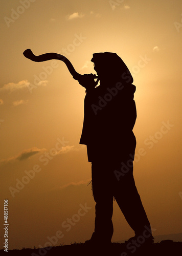 Silhouette of the entire body of a Jewish man wearing a tallit and tefillin and blowing a long shofar made from the horn of a kudu antelope at sunrise during morning prayers  in Israel. photo