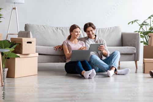 Glad smiling caucasian husband and wife in casual planning new interior with tablet and computer in living room