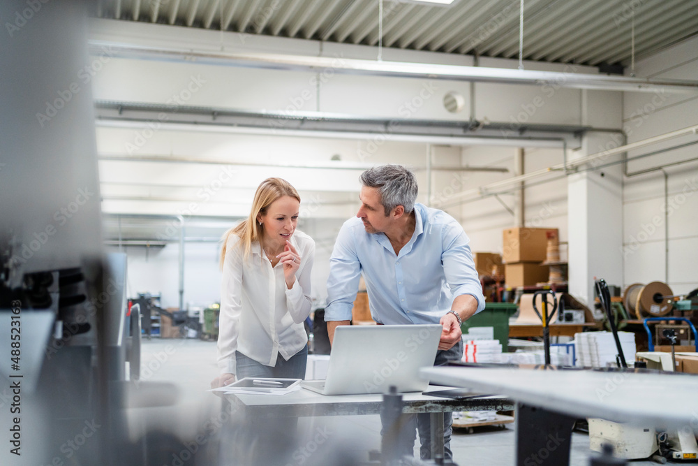Businessman looking at colleague discussing ideas at desk in factory Businessman looking at colleague discussing ideas at desk in factory