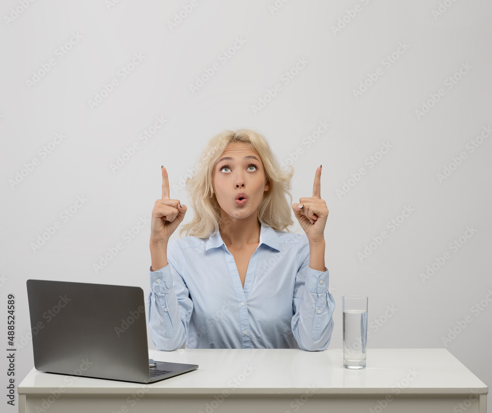 Surprised female office worker sitting at her desk with laptop, looking and pointing upwards at free space
