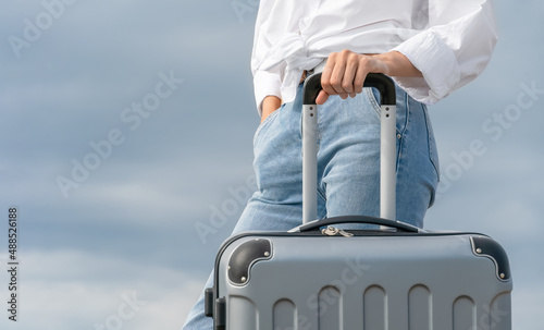 Close-up of a woman's hand holding the handle of a gray suitcase against a gray sky with clouds, copy space