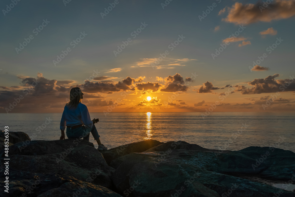 Young woman in a white shirt and jeans is sitting on huge rocks and looking at a beautiful, mesmerizing sunset over the sea, photo from the back, copy space