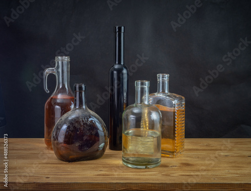 There are five old glass bottles of different shapes and colors with alcoholic beverages  on a wooden table against a background of dark drapery. Bokeh effect.