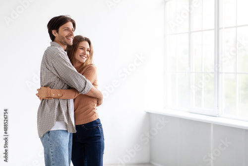 Cheerful millennial caucasian male and female hugging, looking at window in empty room of new apartment, sun flare