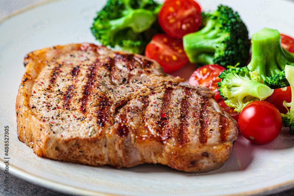 Grilled pork steak with broccoli and tomatoes on plate, dark background. Grilled pork steak with broccoli and tomatoes on plate, dark background.