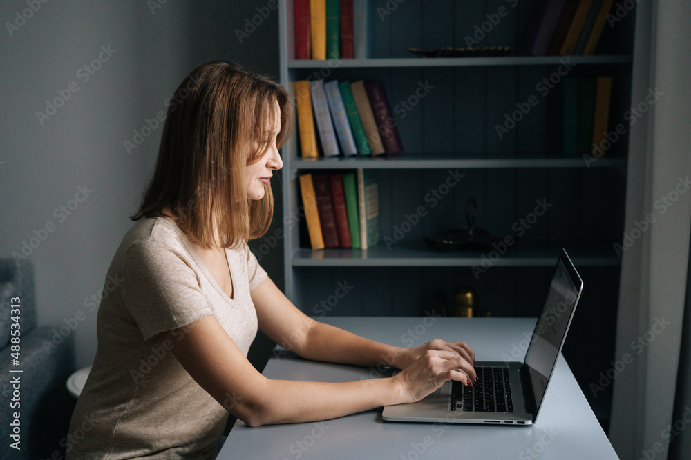 Side view of focused business woman working on laptop computer from home office sitting at desk in dark room. Serious businesswoman studying online, web surfing information, shopping in internet store Side view of focused business woman working on laptop computer from home office sitting at desk in dark room. Serious businesswoman studying online, web surfing information, shopping in internet store
