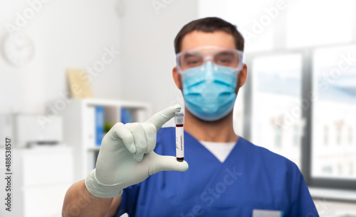 healthcare  coronavirus and medicine concept - doctor in blue uniform  face protective medical mask  goggles and gloves with blood in test tube over medical office at hospital background