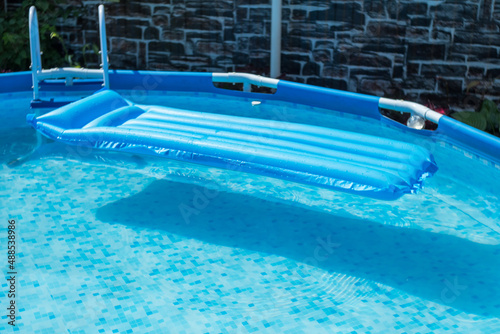 Blue inflatable mattress in a round frame pool of a country house. The pool has the purest blue water. In the background is a dark stone wall.