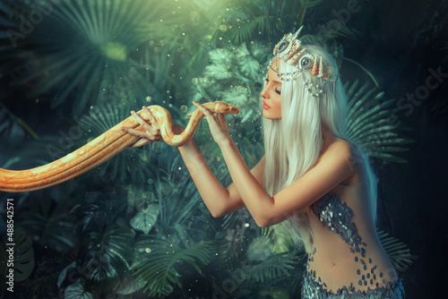 Legend mythical Nagga woman queen sea touch holds milk snake in hands. Blond girl long white flowing hair. Backdrop landscape tropical plants, green leaves. Girl mermaid silver color sexy art costume. photo