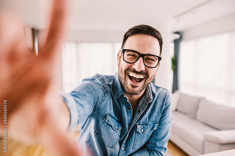 Obraz premium A laughing man grabbing camera and taking a selfie at his new cozy home.