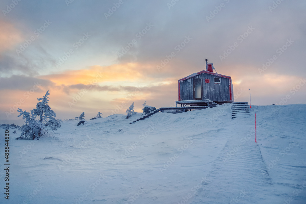 Cabin in winter dawn. Snow-covered staircase leading through snowdrifts to a lonely house on a hilltop in the cool evening.
