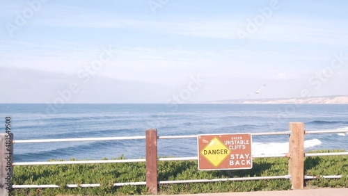 Ocean waves crashing on beach or bluff, La Jolla shore waterfront promenade, California USA. Succulent green ice plant, pacific coast. Seascape view and railings. Danger unstable cliff warning sign.