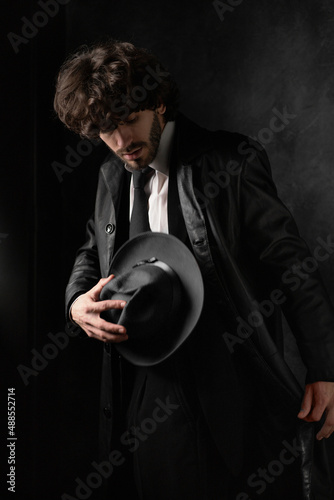 Young man with beard in white shirt and black leather coat with hat against black background. High quality photo