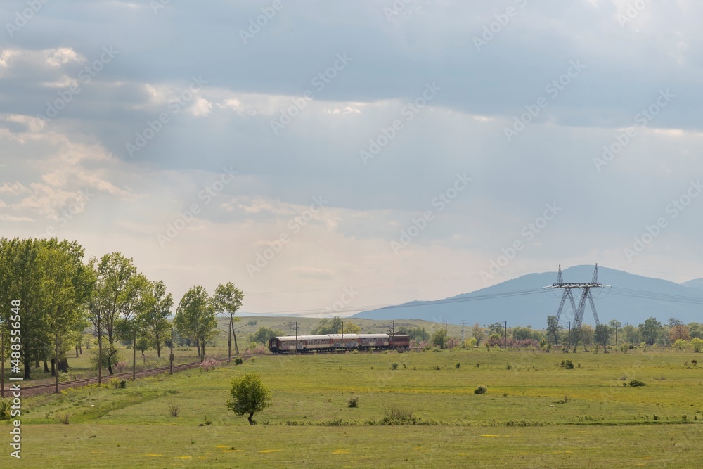 Beautiful landscape, train rides through green field and trees, sun rays break through the clouds and dark blue sky, Bulgaria