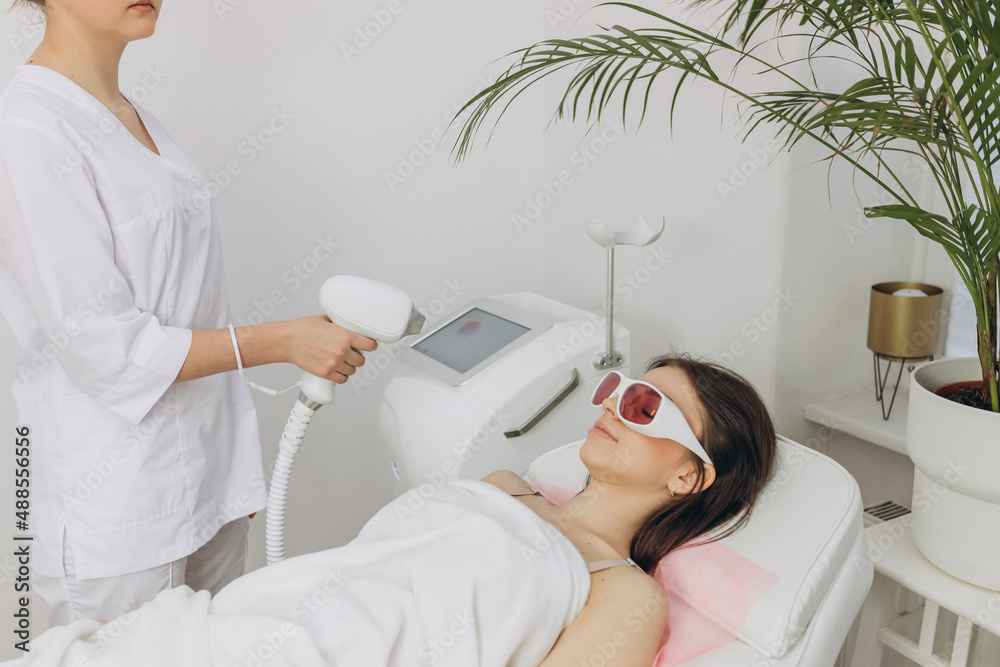 A woman tunes a laser hair removal machine. Girl holding a working part of the epilator in a modern beauty salon. Hair removal, laser procedure at medical clinic, spa concept. Laser skin care banner A woman tunes a laser hair removal machine. Girl holding a working part of the epilator in a modern beauty salon. Hair removal, laser procedure at medical clinic, spa concept. Laser skin care banner