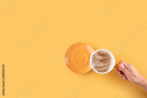 hand holding cup of cappuchino coffee on a yellow background. Space for text.