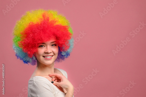 Smiling young attractive female in colorful wig looks at camera on pink background. Close-up.
