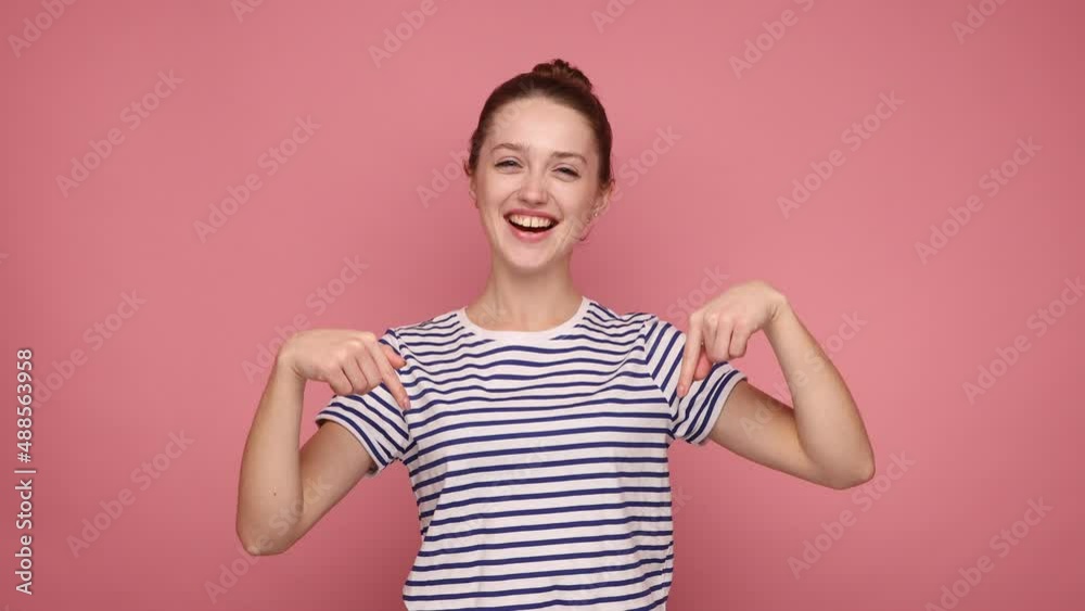 Look, advertise below. Woman with hair bun pointing down place for commercial idea, looking at camera with toothy smile, wearing striped T-shirt. Indoor studio shot isolated on pink background. Look, advertise below. Woman with hair bun pointing down place for commercial idea, looking at camera with toothy smile, wearing striped T-shirt. Indoor studio shot isolated on pink background.