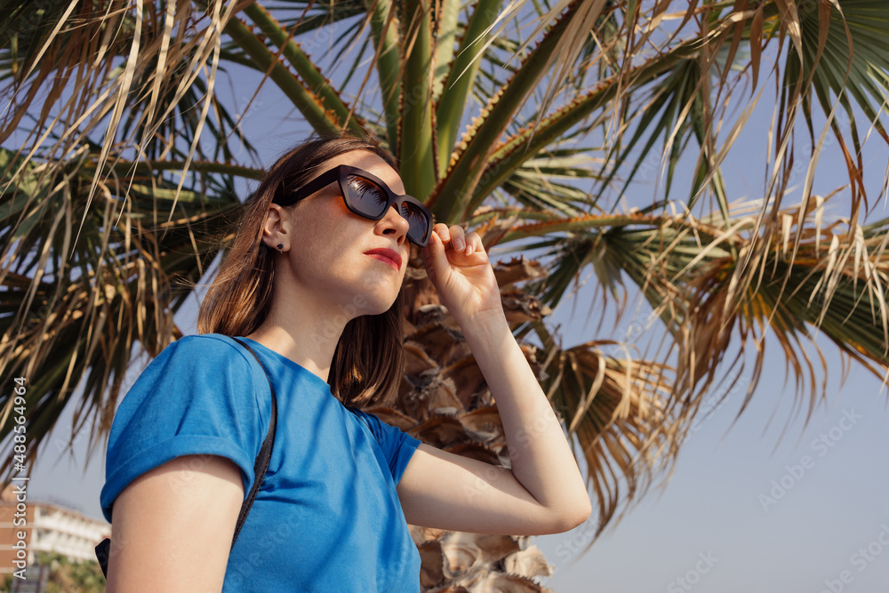 A woman in a blue t shirt and brown sunglasses enjoy the view of the sea with a palm tree in background