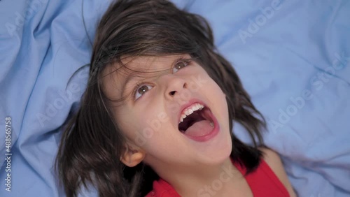 joyful caucasian child is a dark haired boy 5 years with long hair and a red T-shirt lying on the bed on his back