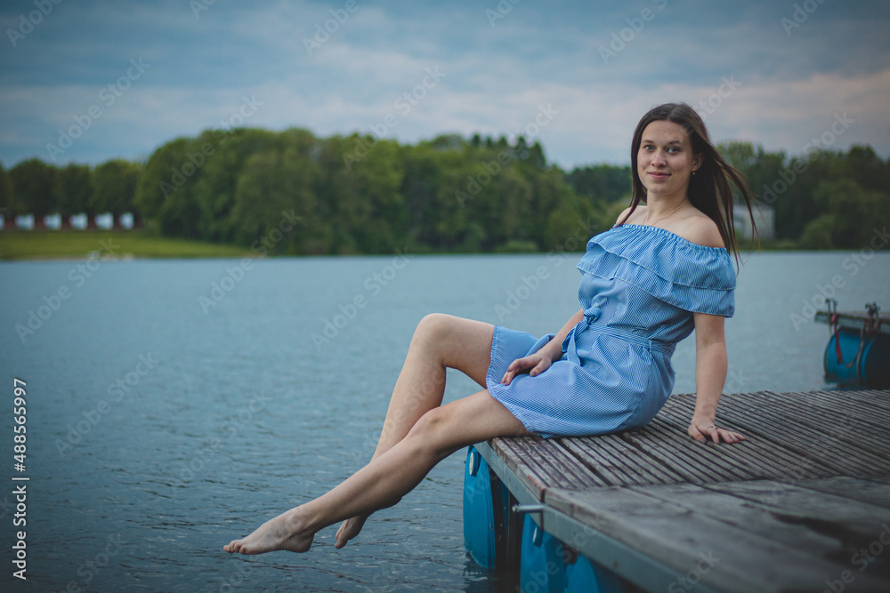 Cute brunette model with long straight dark hair in a summer blue dress with a beautiful realistic smile poses by a lake in the Czech Republic. European woman. Candid portrait with realistic smile