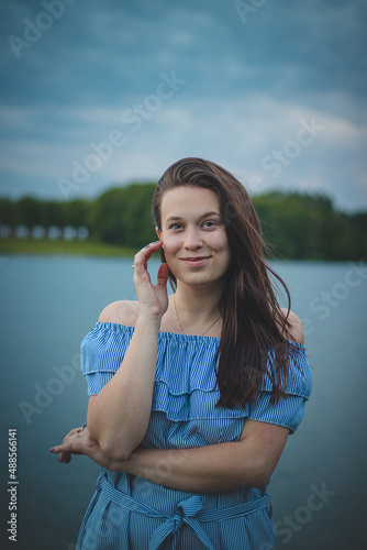 Cute brunette model with long straight dark hair in a summer blue dress with a beautiful realistic smile poses by a lake in the Czech Republic. European woman. Candid portrait with realistic smile © Fauren