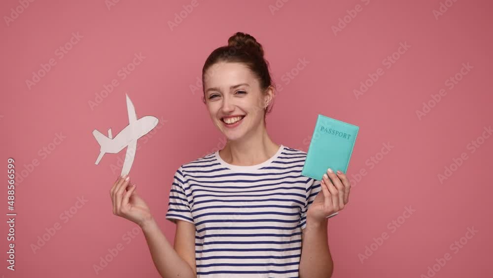 Extremely happy excited woman with bun hairstyle holding paper plane and passport, being glad she has a vacation, wearing striped T-shirt. Indoor studio shot isolated on pink background. Extremely happy excited woman with bun hairstyle holding paper plane and passport, being glad she has a vacation, wearing striped T-shirt. Indoor studio shot isolated on pink background.