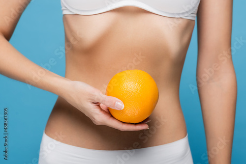 cropped view of young woman in white bra holding ripe orange isolated on blue