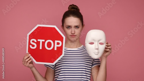 Woman holding white mask and stop traffic sign to camera, nodding her head no, don't change your personality, wearing striped T-shirt. Indoor studio shot isolated on pink background. photo
