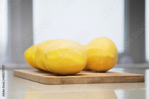 Several peeled potatoes on a wooden cutting board on a white table.