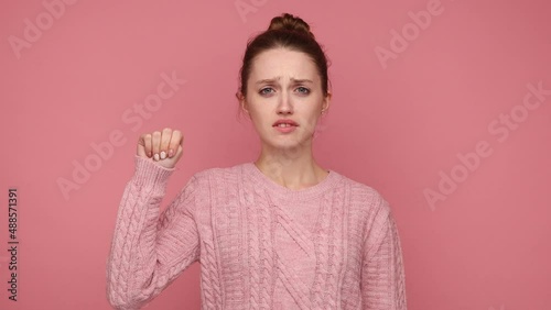Sad scared woman with hair bun showing four fingers folding over the thumb, the violence at home, signal for help, wearing knitted sweater. Indoor studio shot isolated on pink background.