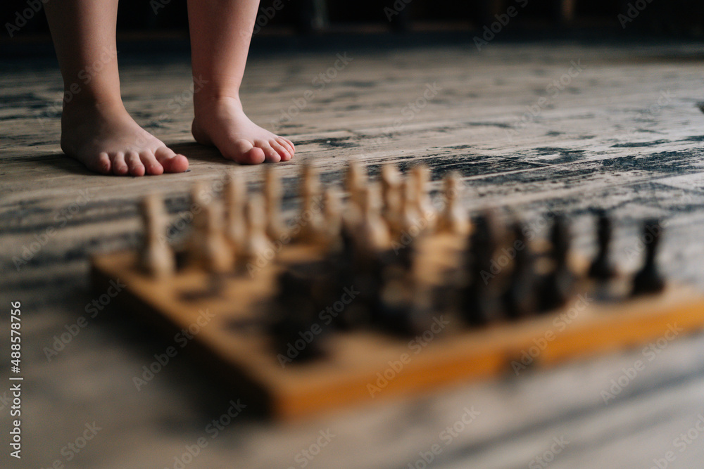Cropped selective focus shot of barefoot legs unrecognizable woman standing near chessboard on wooden floor in dark room, thinking chess move. Intelligent lady playing logical game alone at home