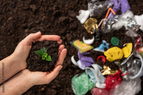 Hands holding small plant on a blurred background of black soil and plastic waste