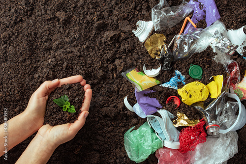 Hands holding a young sprout against the ground polluted with plastic trash