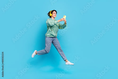 Full length photo of funky millennial brunette lady run look telephone wear headphones pullover jeans shoes isolated on blue background